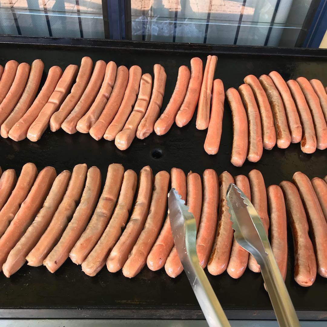 Rows of hot dogs cooking on a black griddle, with metal tongs in the foreground.