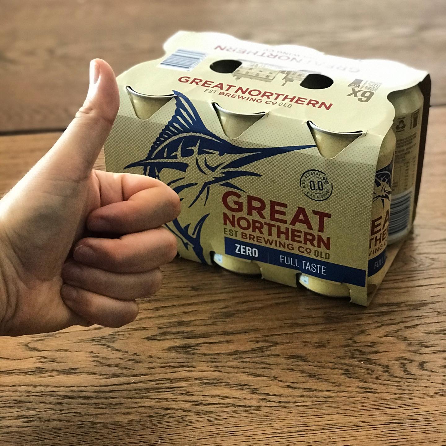 Hand giving a thumbs-up beside a six-pack of Great Northern Zero cans on a wooden table.