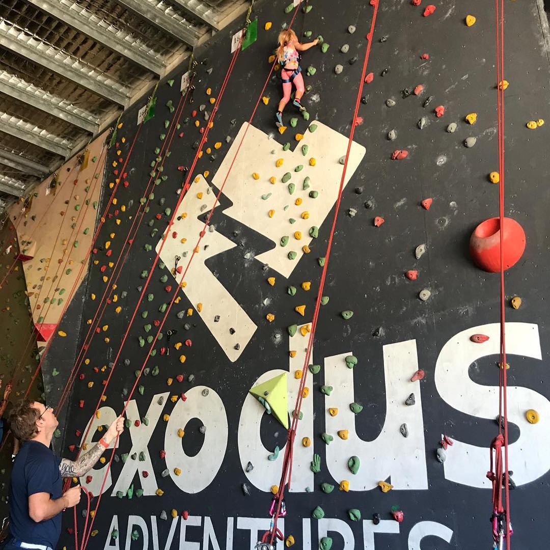 Child climbs an indoor rock wall with colorful holds while an adult below manages the rope.