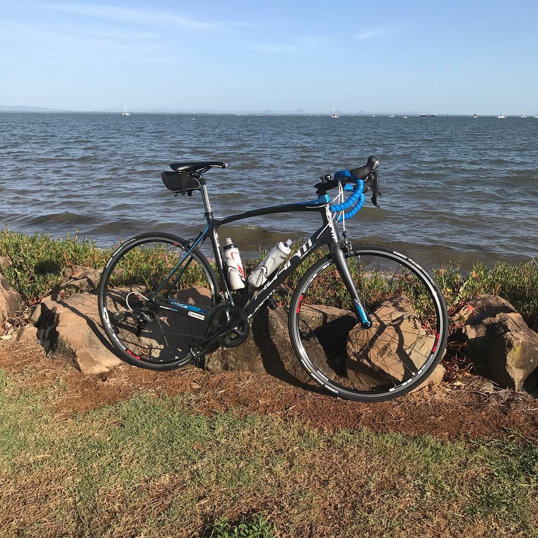 Black road bike with blue handlebar tape leaning on rocks beside a bay, with small boats on the water under a clear blue sky.