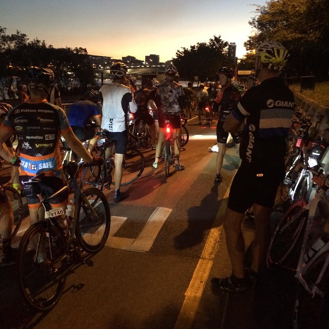 A group of cyclists in helmets stand with their bikes on a road at dawn, with city buildings silhouetted in the distance.
