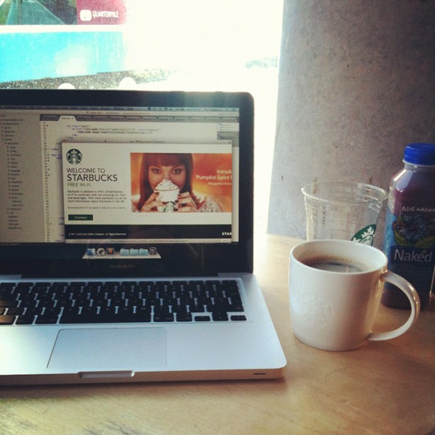 Laptop on a table displaying the Starbucks website beside a white coffee mug, a clear plastic cup, and a bottle of Naked juice.