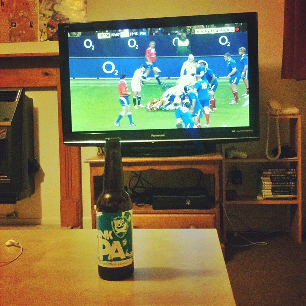 Beer bottle on a coffee table in front of a TV showing a rugby match in a living room.