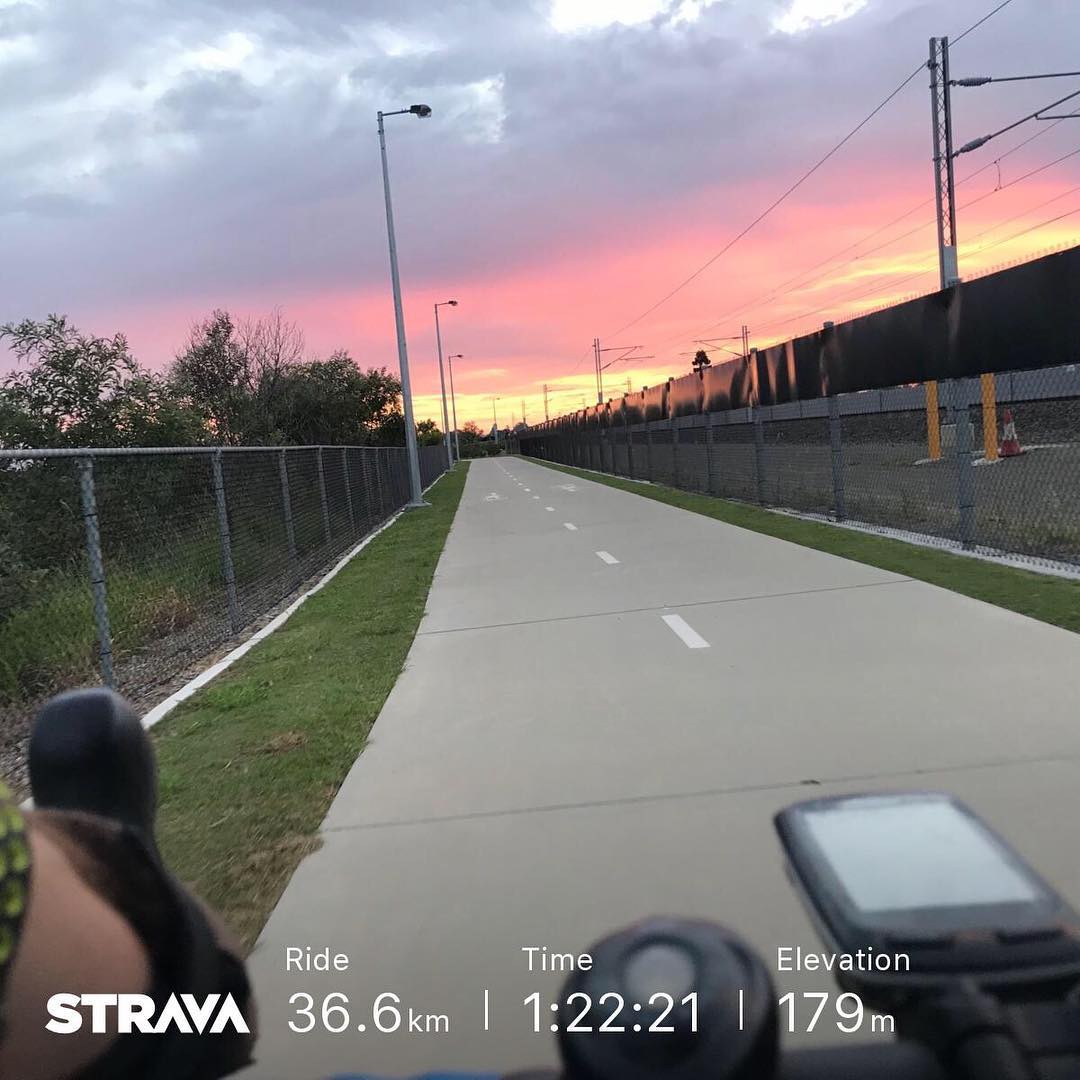 Paved bike path between chain-link fences at sunset, with pink clouds, streetlights, rail lines, and bike handlebars with a screen in front.