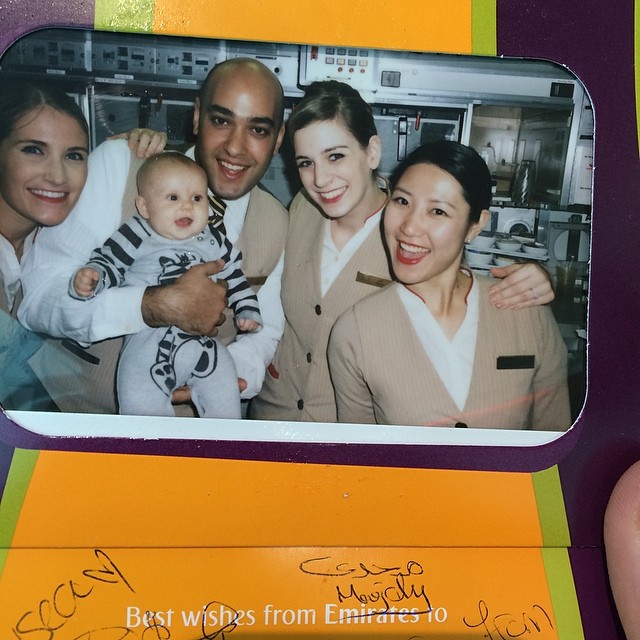 Five smiling adults in uniforms pose with a baby in a striped outfit in a printed photo with signatures below.