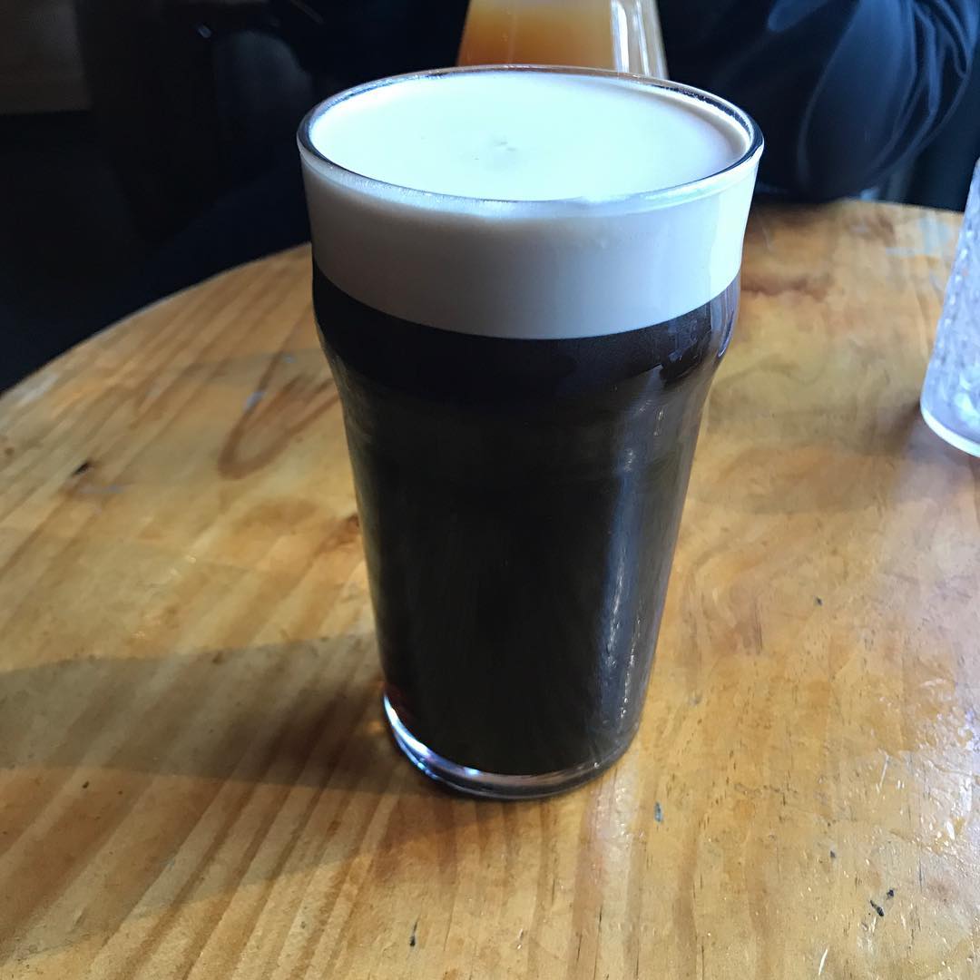 A pint glass of dark beer with a thick creamy foam head on a wooden table.