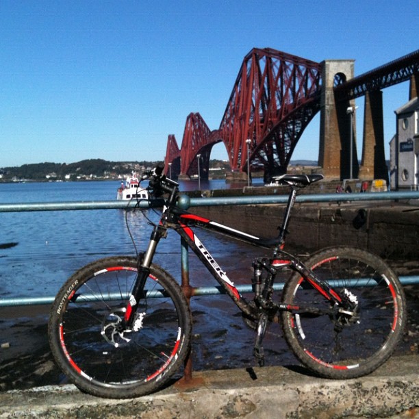 Black and red mountain bike parked by a waterfront railing, with a large red cantilever bridge in the background.