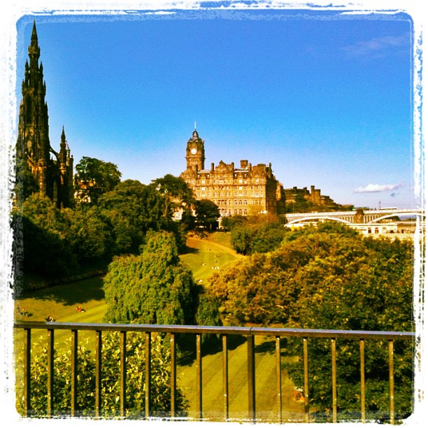 View over a green park with trees, a black railing, a tall dark spire, and a clock-topped stone building under a clear blue sky.
