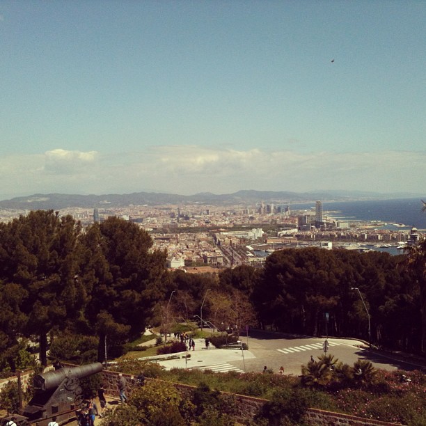 Panoramic city view from a hillside with trees, a road, distant buildings, mountains, and the sea under a clear sky.
