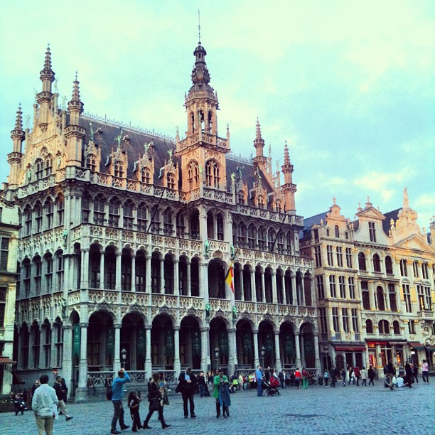Ornate historic building with spires and arched colonnades facing a cobblestone square with scattered pedestrians.