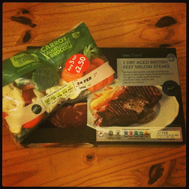 Bag of mixed vegetables and a Tesco Finest two-pack of dry aged British beef sirloin steaks on a wooden table.