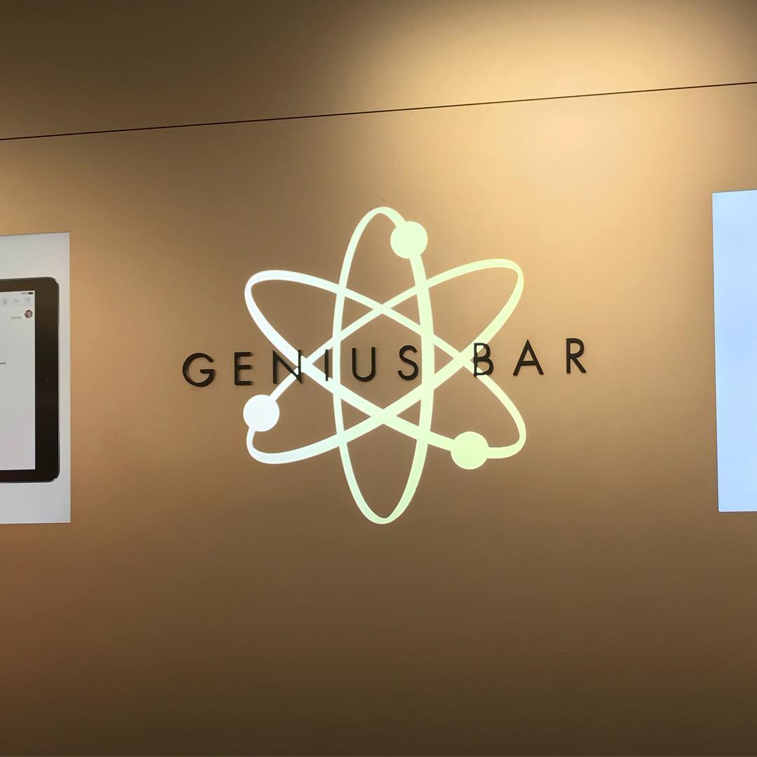 Glowing atom symbol over the words “GENIUS BAR” on a tan wall, with partial screens visible at both sides.
