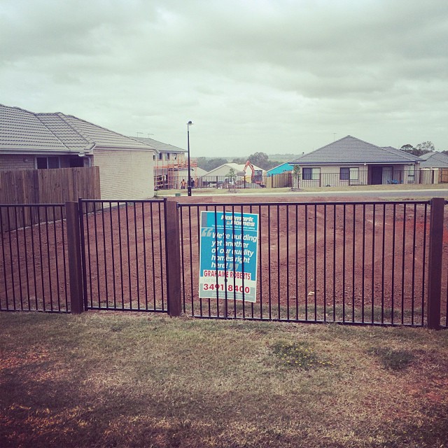 Black metal fence around an empty dirt lot with a blue real estate sign, houses, and cloudy sky in the background.