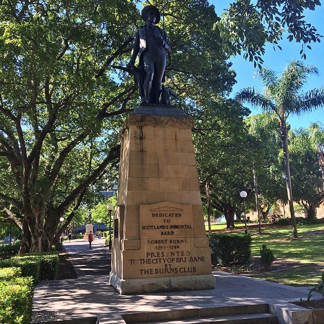 Statue of Robert Burns on a tall stone pedestal in a leafy park with paths, trees, and a clear blue sky.