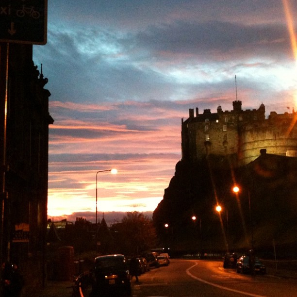 Sunset sky over a curved city street with parked cars, streetlights, and a castle-like building on a hill in silhouette.