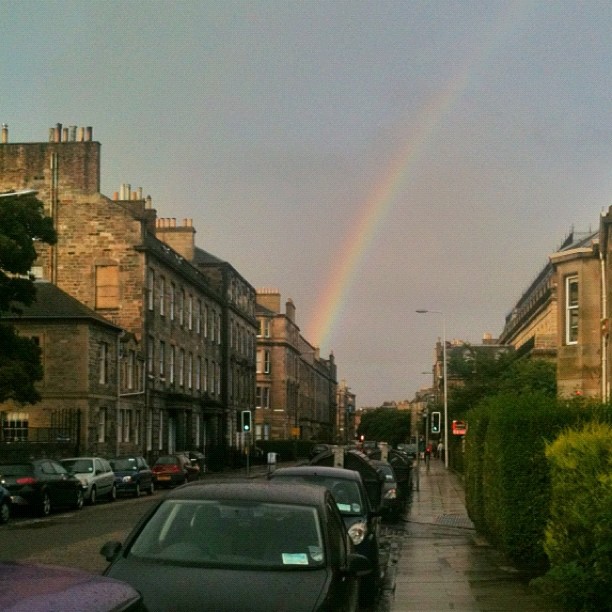 Rainbow over a wet city street lined with parked cars, stone buildings, and hedges.