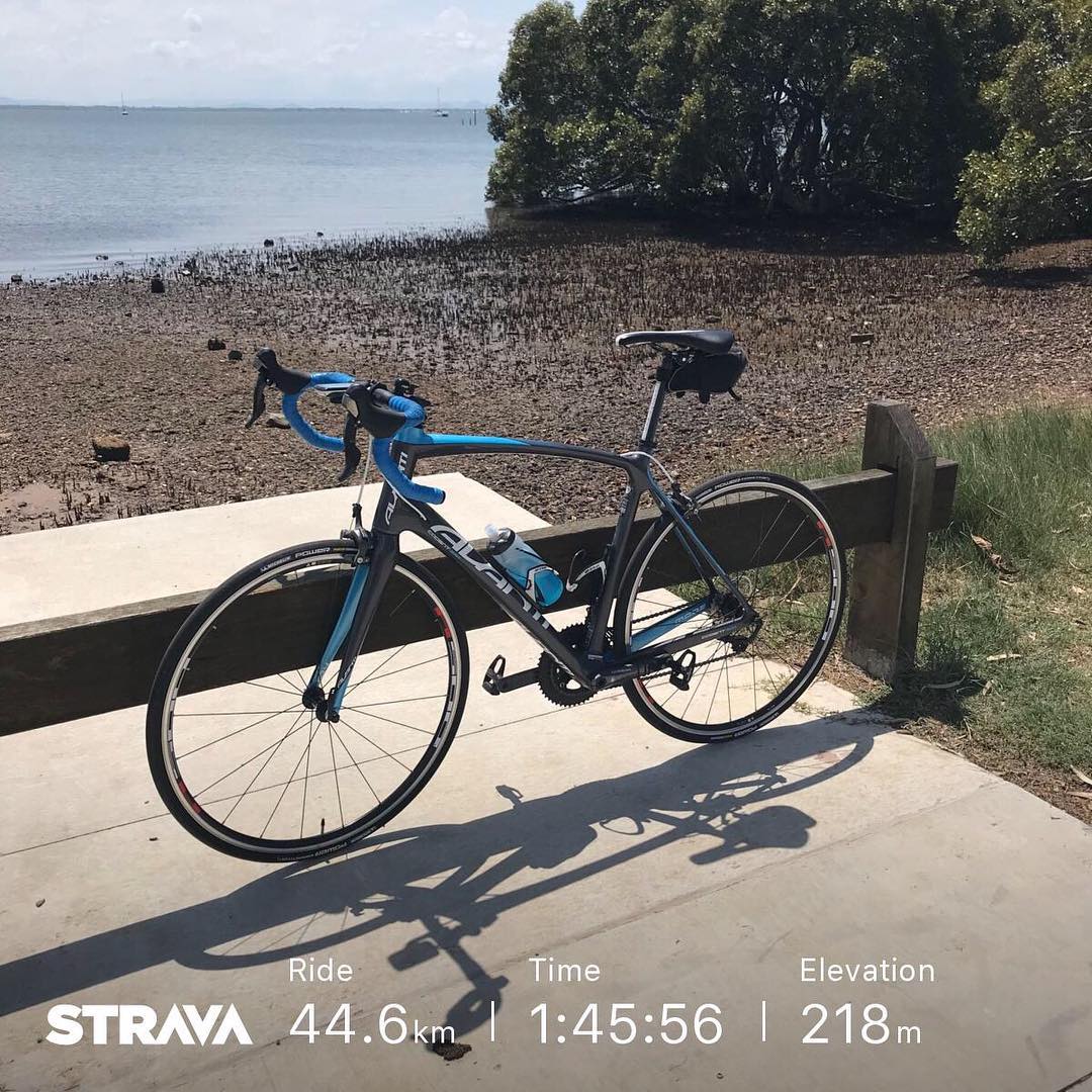 Blue and black road bike on a paved path by a muddy shoreline and calm water, with Strava stats overlaid at the bottom.