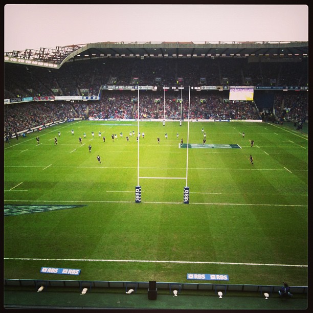 Wide view of a rugby match in a large stadium, with players spread across the field and goalposts centered in the foreground.