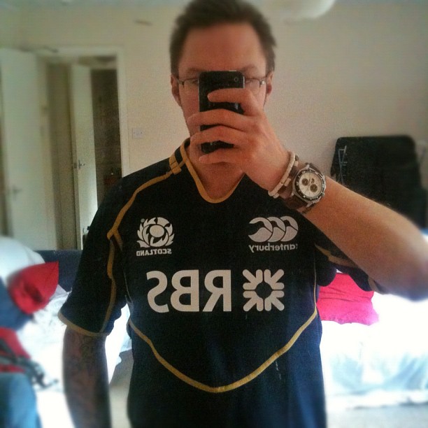 Person taking a mirror selfie in a bedroom, wearing a black Scotland rugby jersey and a large wristwatch.