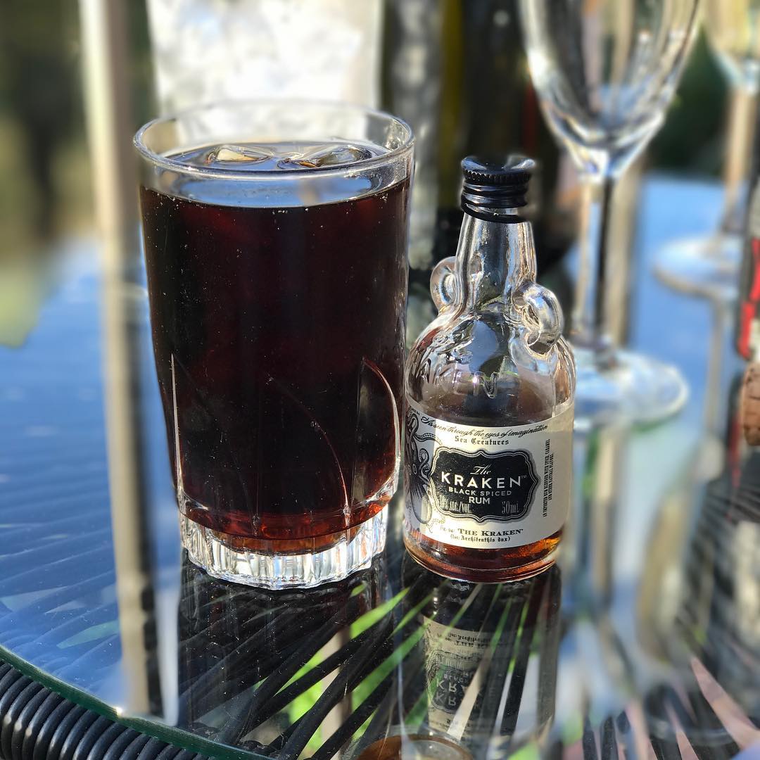 Glass of dark iced drink beside a small bottle of Kraken Black Spiced Rum on a reflective table.
