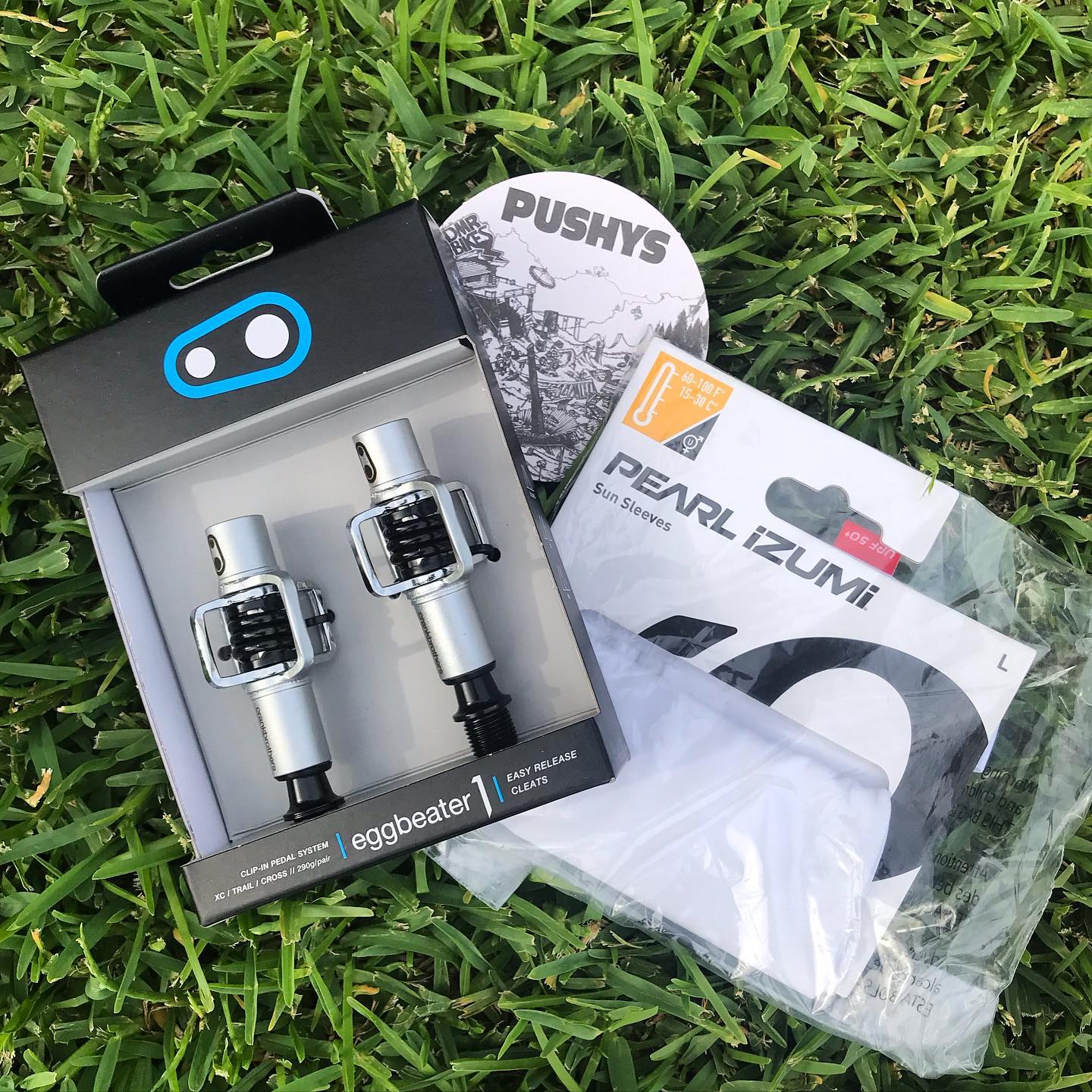 Boxed clip-in bike pedals, white Pearl Izumi sun sleeves in packaging, and a round sticker placed on green grass.