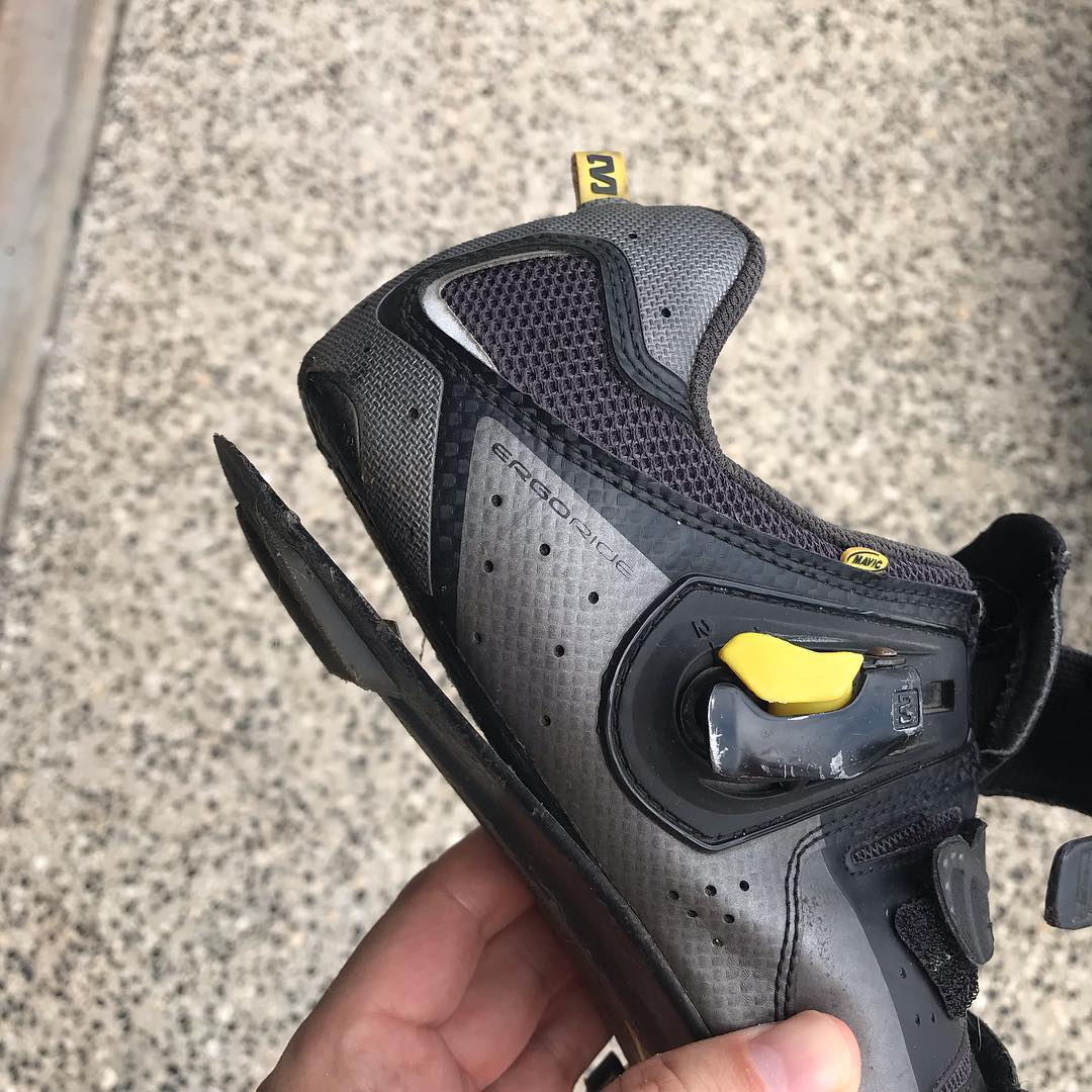 Black and gray cycling shoe with the sole peeling away at the heel, held over a speckled stone surface.