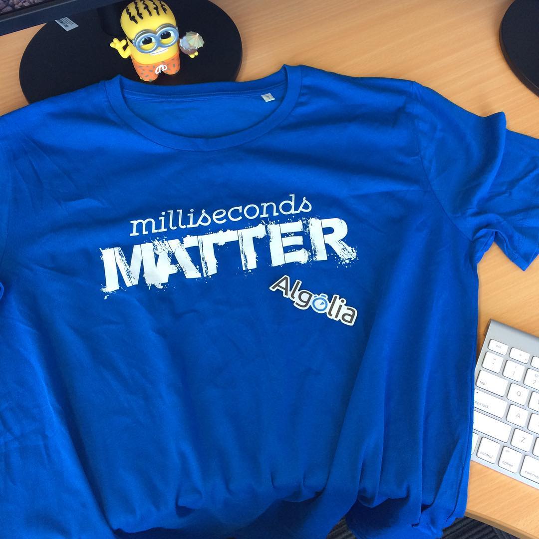 Blue T-shirt on a desk with “milliseconds MATTER Algolia” printed in white, next to part of a keyboard and a small Minion figure.