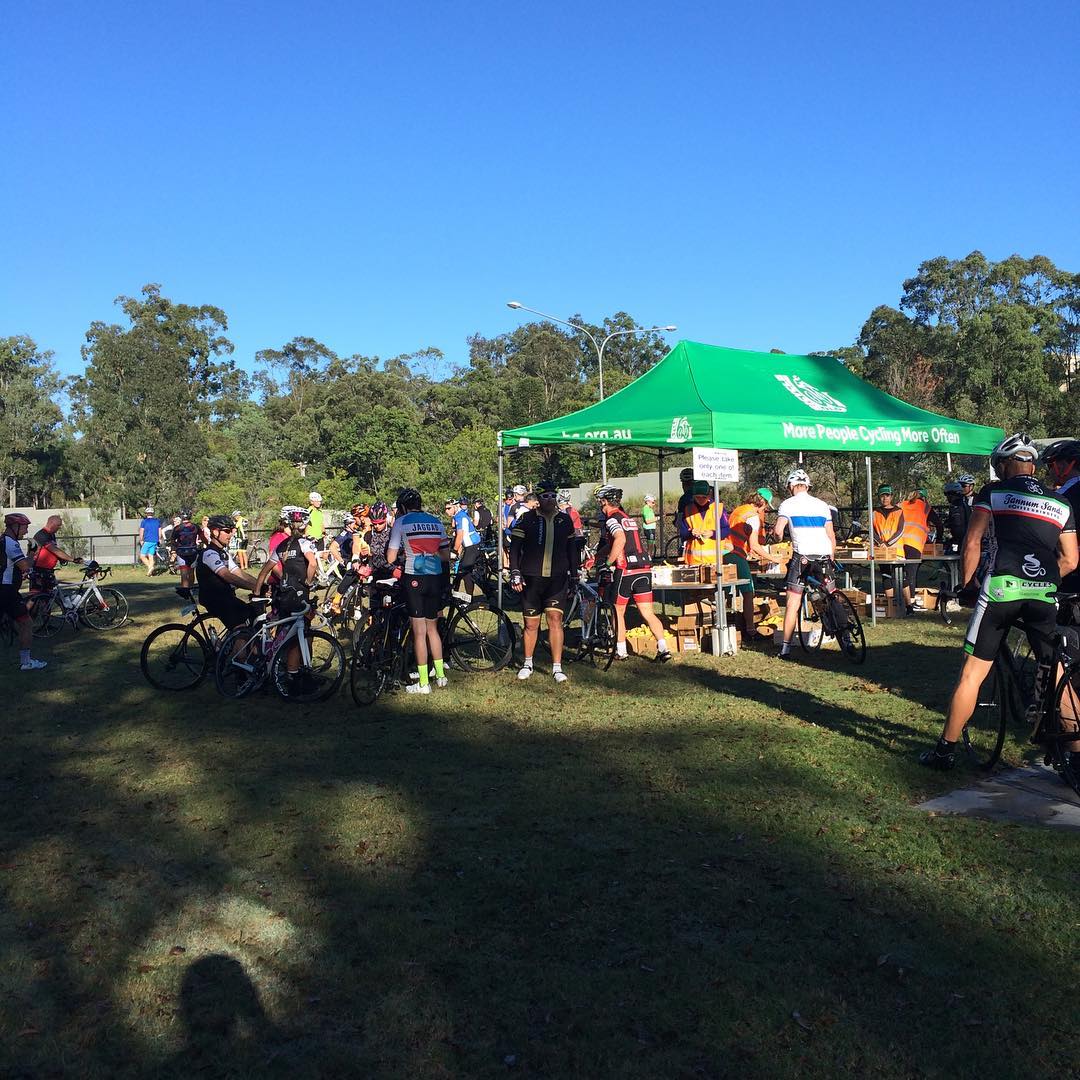 Cyclists gather with their bikes around a green tent in a grassy park under a clear blue sky.