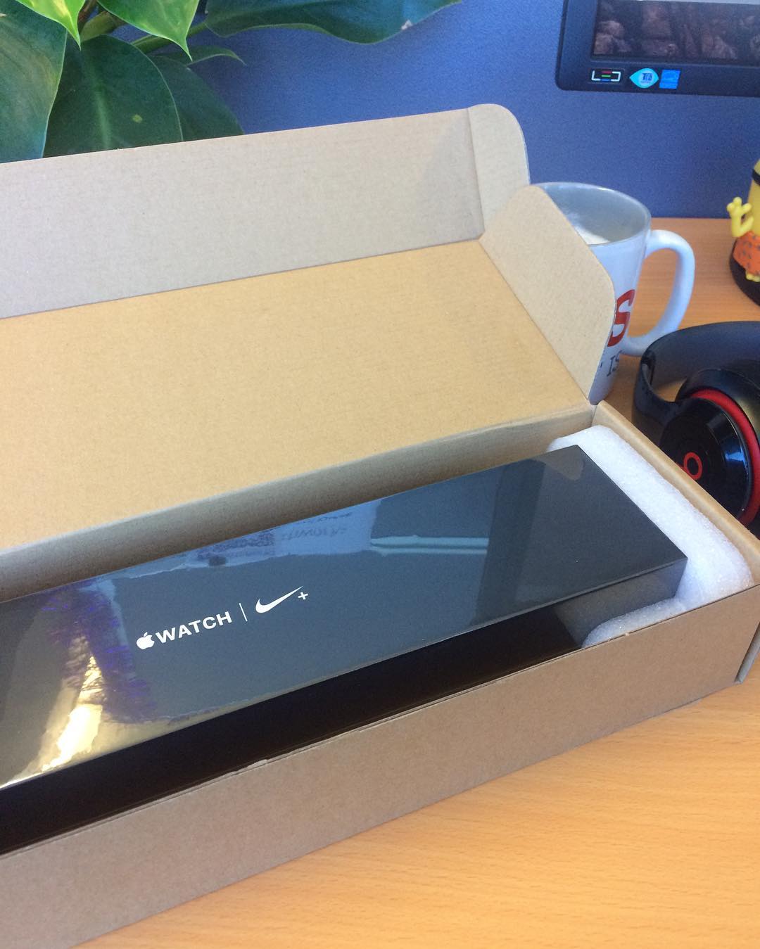 Open cardboard box on a desk containing an Apple Watch Nike+ package, with a mug, headphones, and plant nearby.