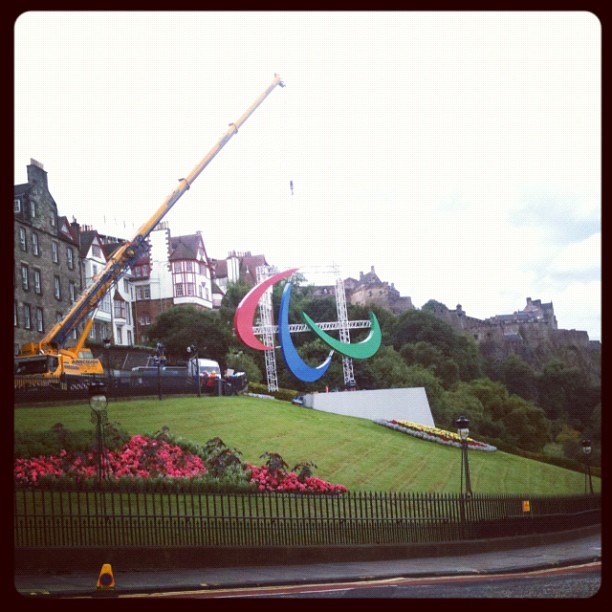 A crane beside a grassy hillside installs large red, blue, and green curved structures near buildings and a castle on a hill.