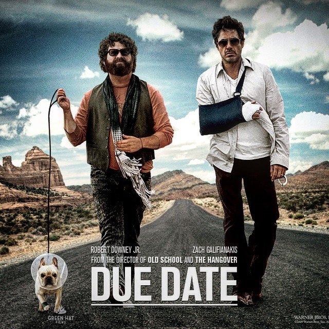 Two men walk down a desert road, one with his arm in a sling and the other holding a leash attached to a small dog in a cone.