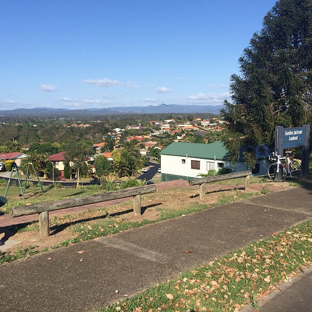 Overlook with a paved path, wooden barrier, large tree, bicycle, and a view of houses and distant mountains under a clear blue sky.