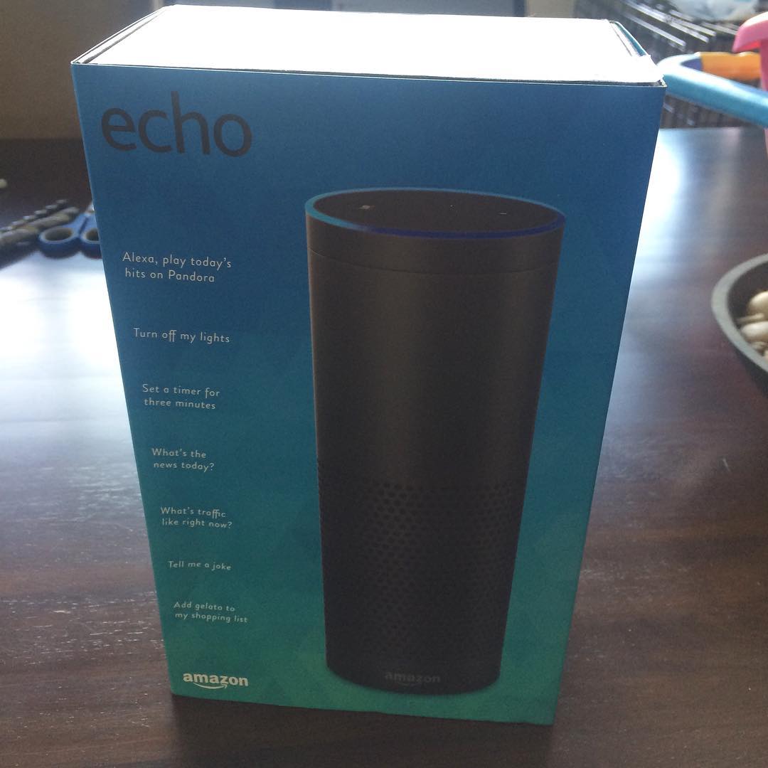 Amazon Echo box standing on a dark table, with the speaker pictured on blue packaging and sample Alexa commands listed on the side.