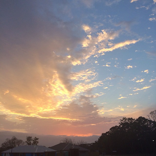 Glowing sunset sky with golden clouds, blue patches, and dark tree silhouettes above rooftops.
