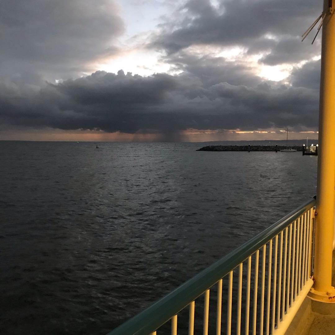 Dark storm clouds over the ocean at dusk, with rain in the distance and a lit railing beside a pier on the right.