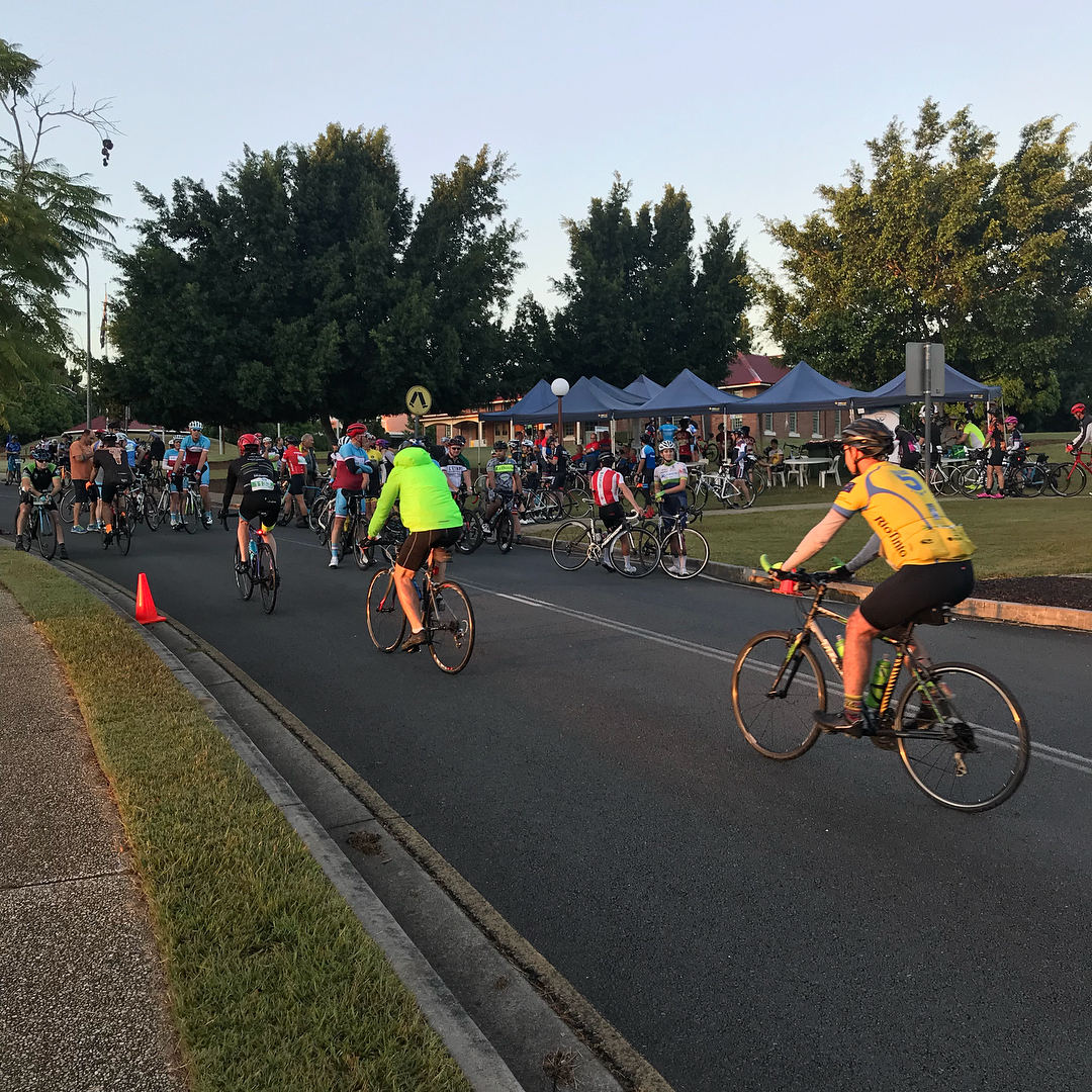 Cyclists gather and ride along a road beside blue tents and trees in early morning light.