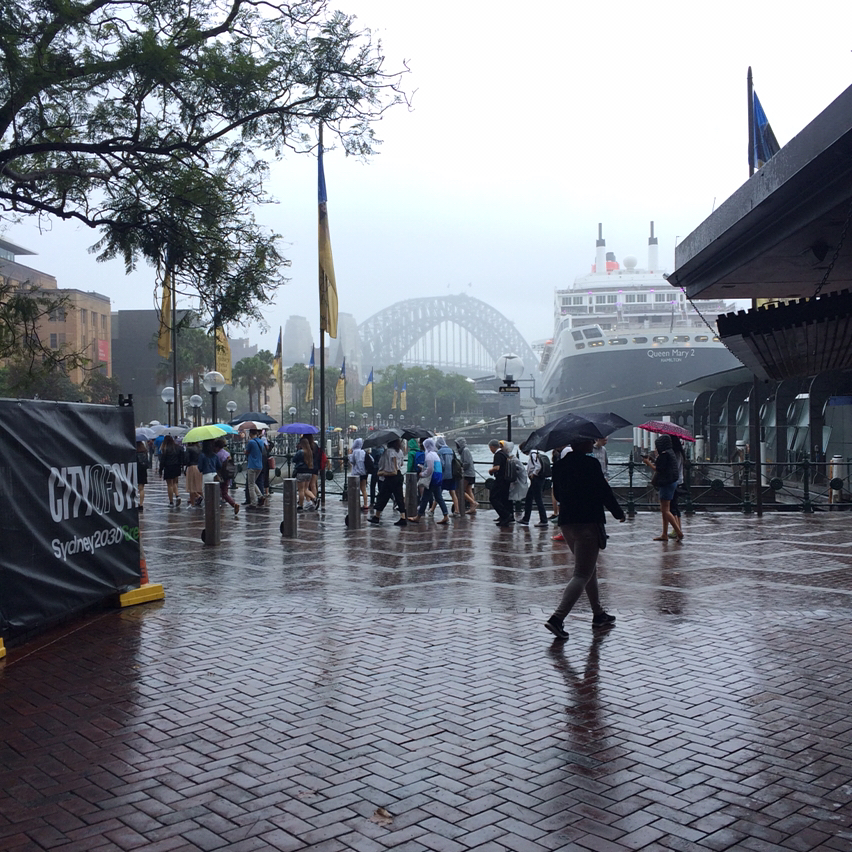 Rainy harbor walkway with pedestrians carrying umbrellas, the Queen Mary 2 docked at right, and the Sydney Harbour Bridge faintly visible in mist.