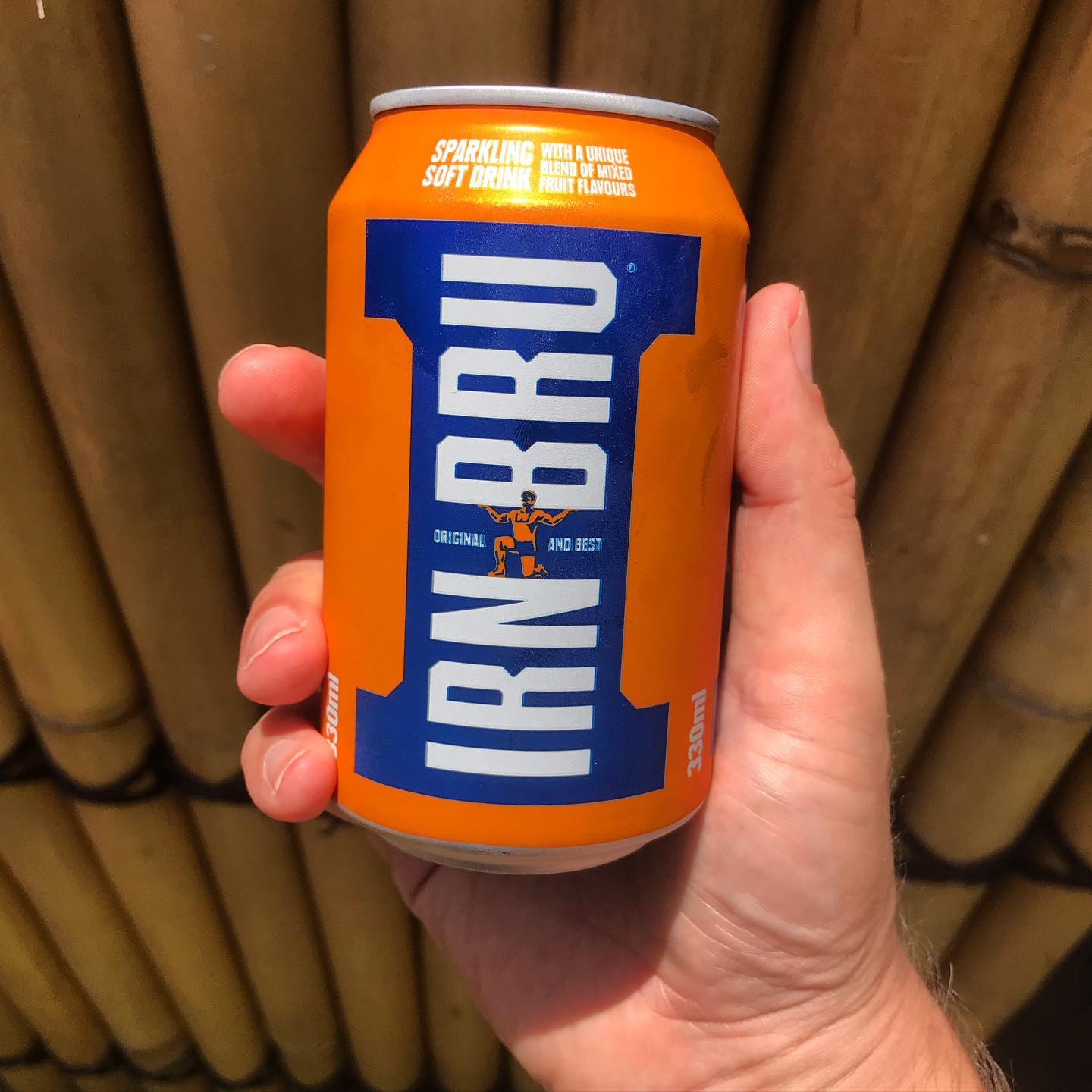 Hand holding an orange can of Irn-Bru sparkling soft drink in front of a bamboo wall.