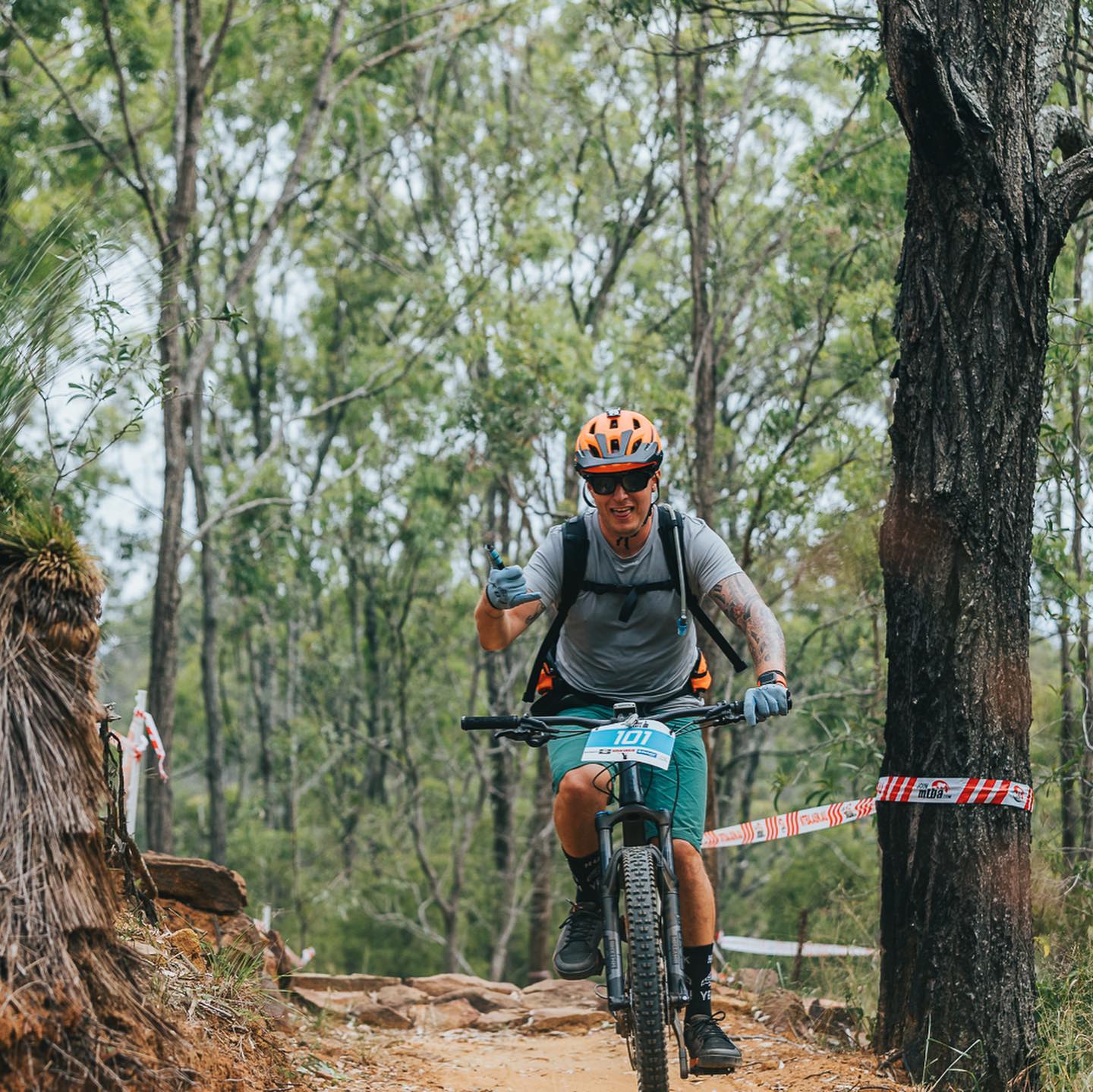 Mountain biker wearing an orange helmet rides a rocky forest trail and gestures with one hand.