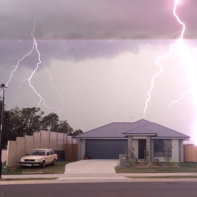 Lightning bolts strike in a stormy sky above a suburban house with a car parked beside a tall fence.
