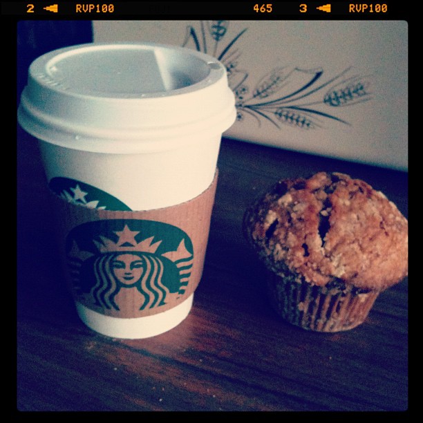 Starbucks takeaway coffee cup beside a crumb-topped muffin on a wooden table.