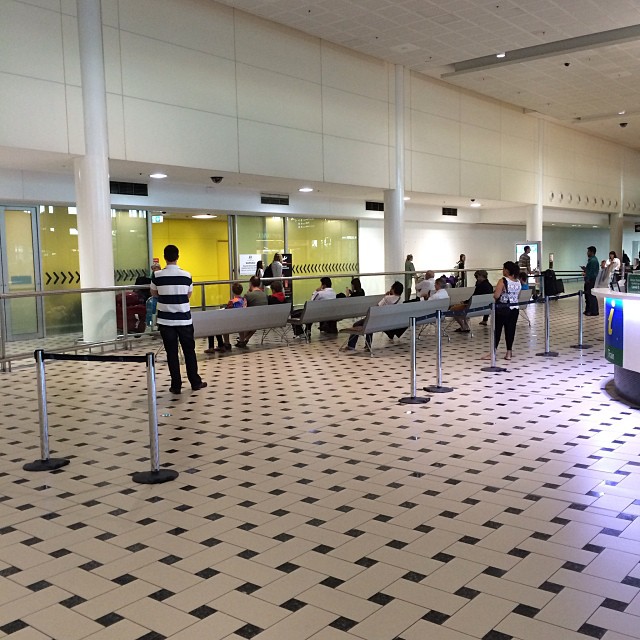 People waiting on benches behind queue barriers in a large tiled indoor hall with columns and yellow-tinted windows.