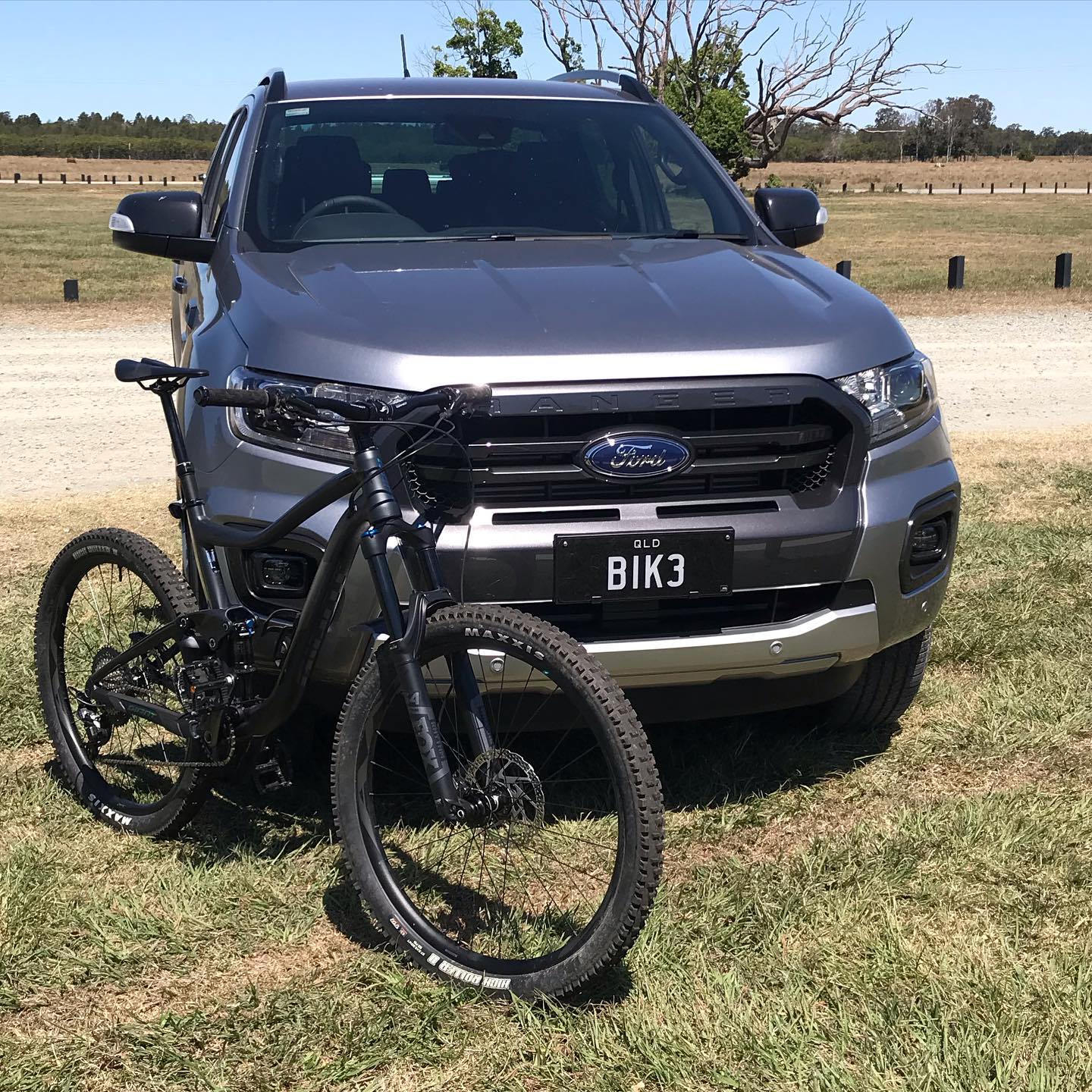 Black mountain bike parked in front of a gray Ford pickup truck on dry grass in an open field.