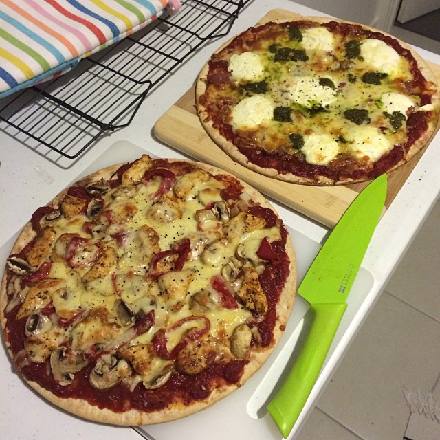 Two homemade pizzas on a counter, one topped with mushrooms and red onion, beside a green knife and cooling rack.