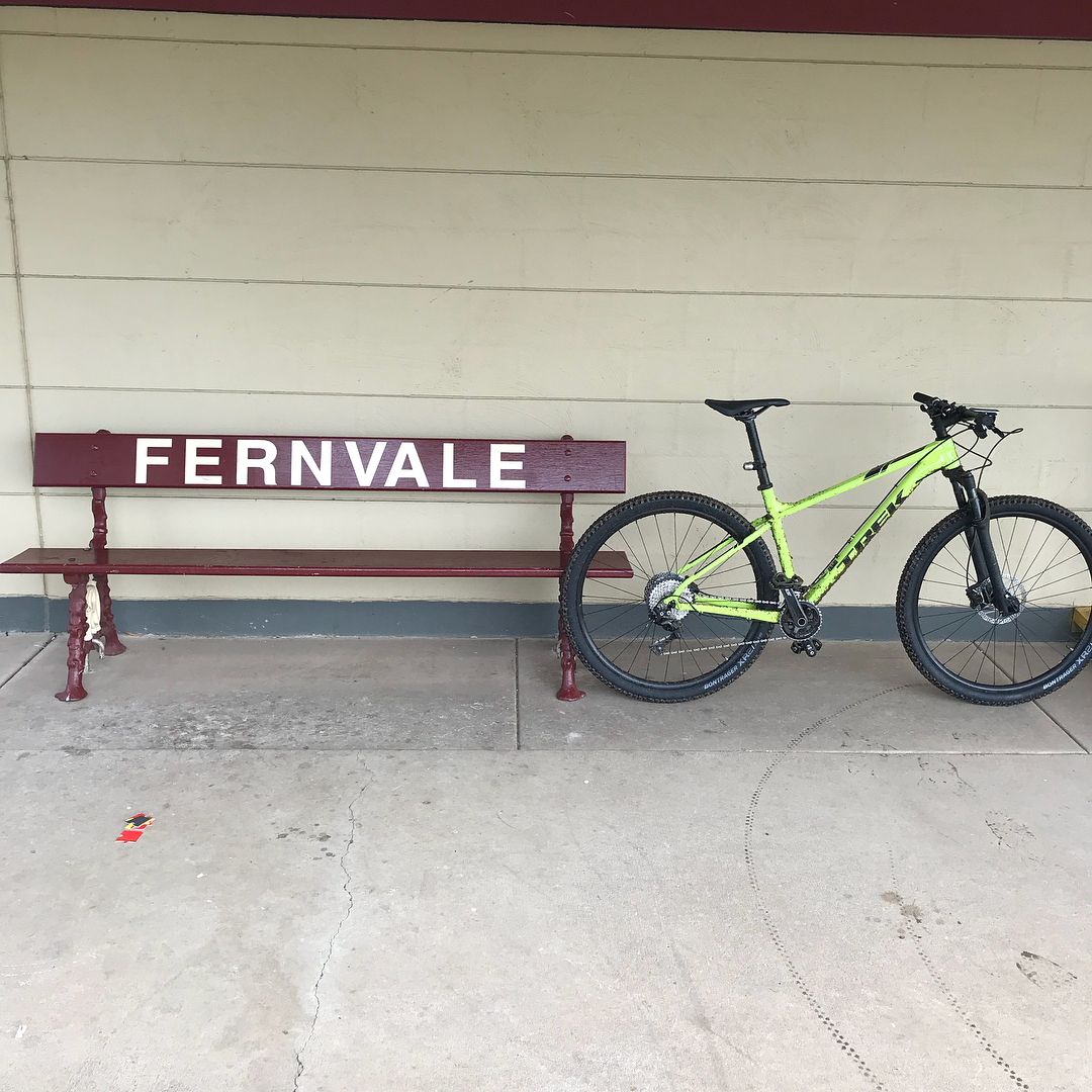 Maroon bench labeled “FERNVALE” beside a bright green mountain bike against a pale wall.