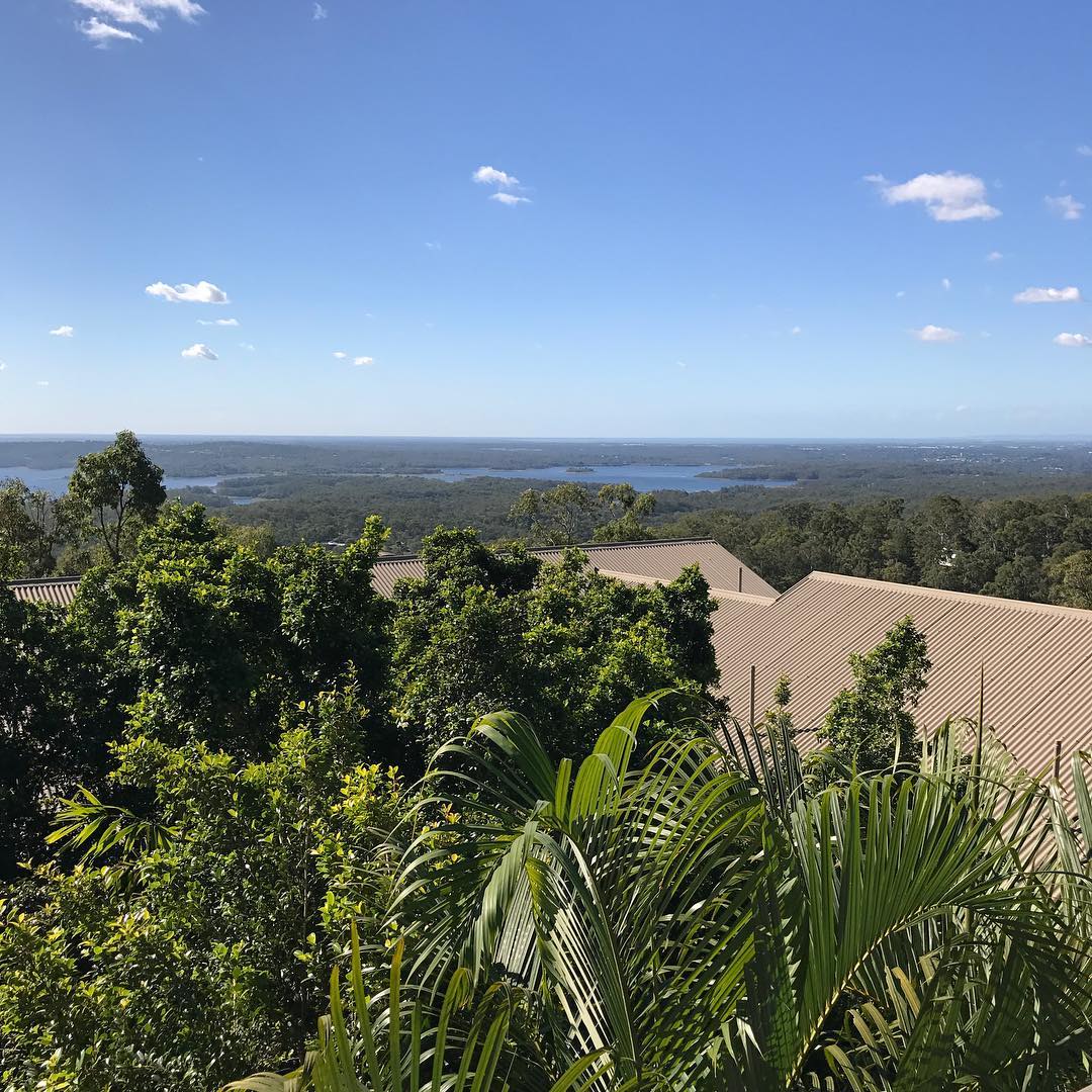 View over dense green trees and rooftops toward a distant lake under a bright blue sky with scattered small clouds.
