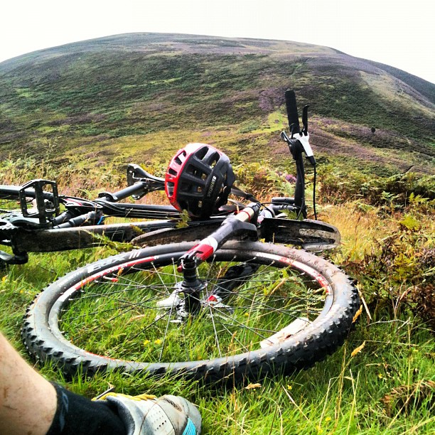 Mountain bike and helmet resting in grassy hillside foreground with a broad, heather-covered hill rising in the background.
