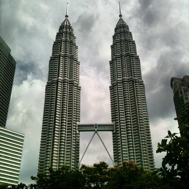 Two tall twin skyscrapers linked by a skybridge rise above trees under a dark cloudy sky.