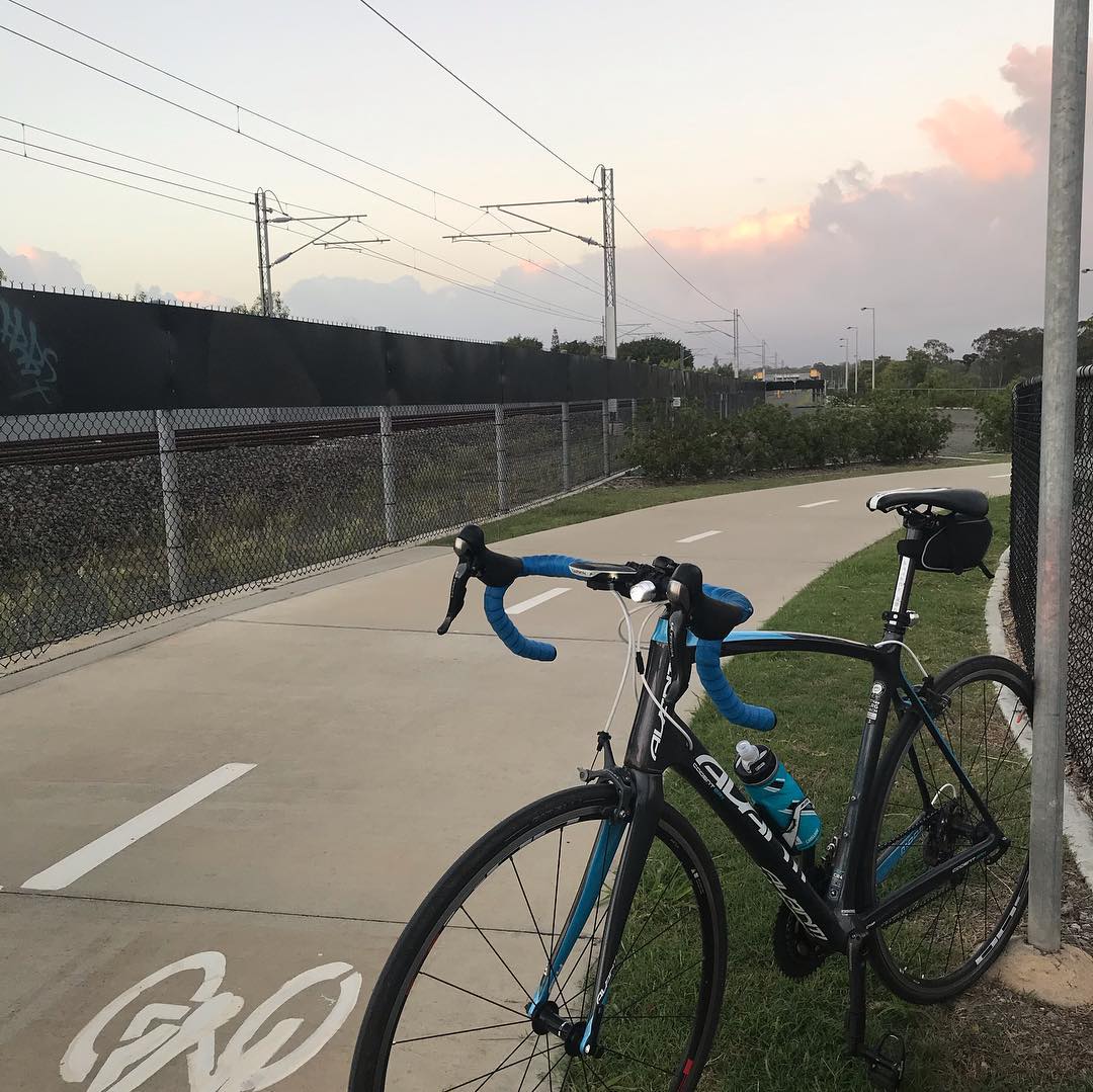 Road bike with blue handlebar tape parked beside a paved path next to railway tracks under a cloudy sunset sky.