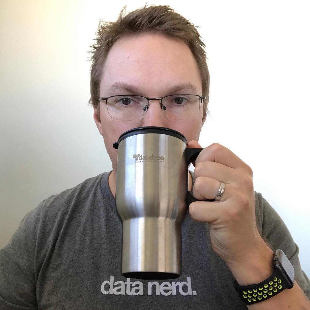 Person wearing glasses and a gray “data nerd.” shirt drinks from a large stainless steel mug, with a smartwatch on one wrist.